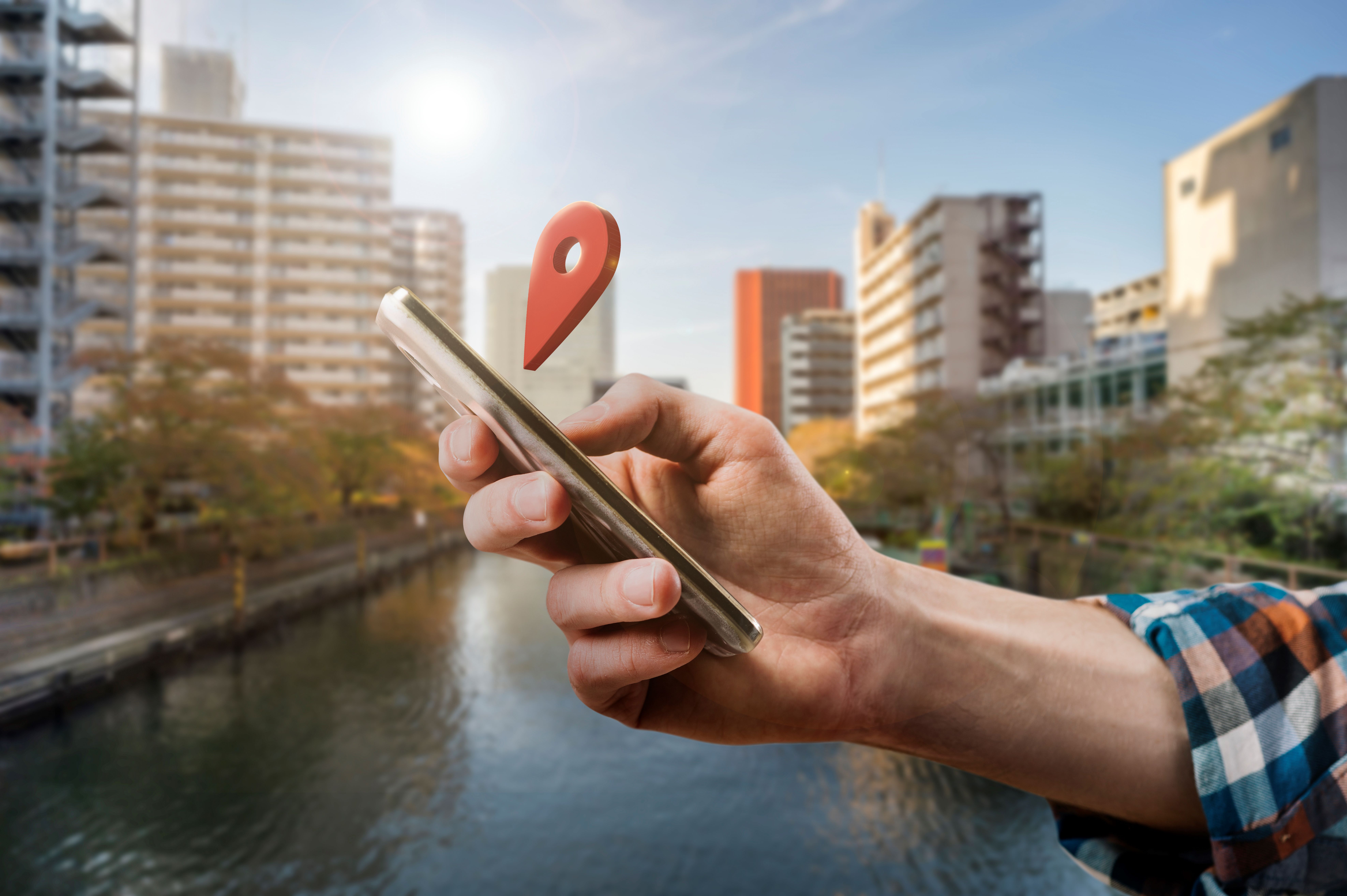 Uma mão segurando um celular sobre uma ponte em um rio na cidade. Acima do celula há um alfinete de local.